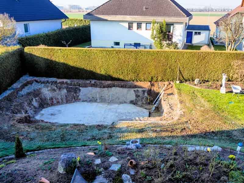 A rectangular hole in a backyard indicating the start of a pool installation, surrounded by grass and adjacent homes.