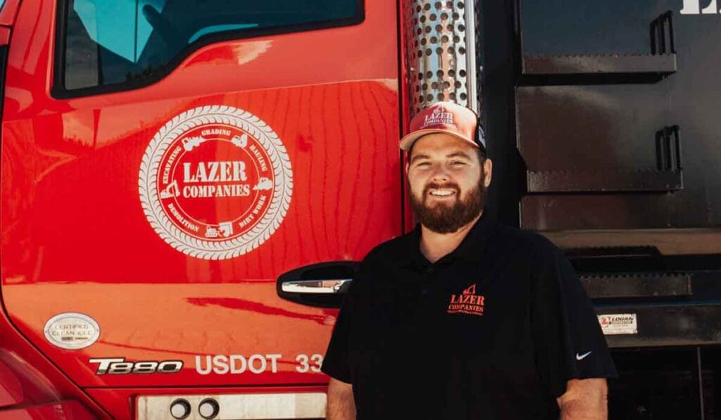 Man standing next to a red truck with "Lazer Companies" logo. He is wearing a black shirt and a baseball cap, both with the same logo.